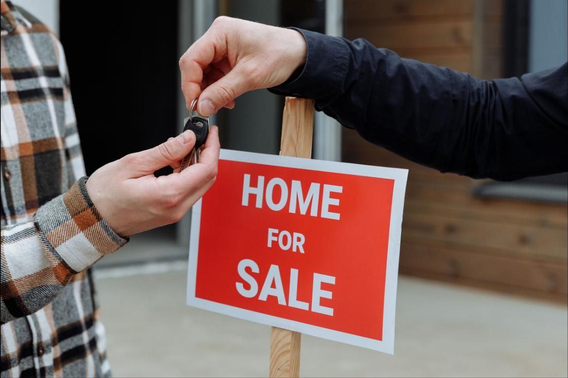 Keys being handed over beside a red and white Home For Sale sign staked in front of a house